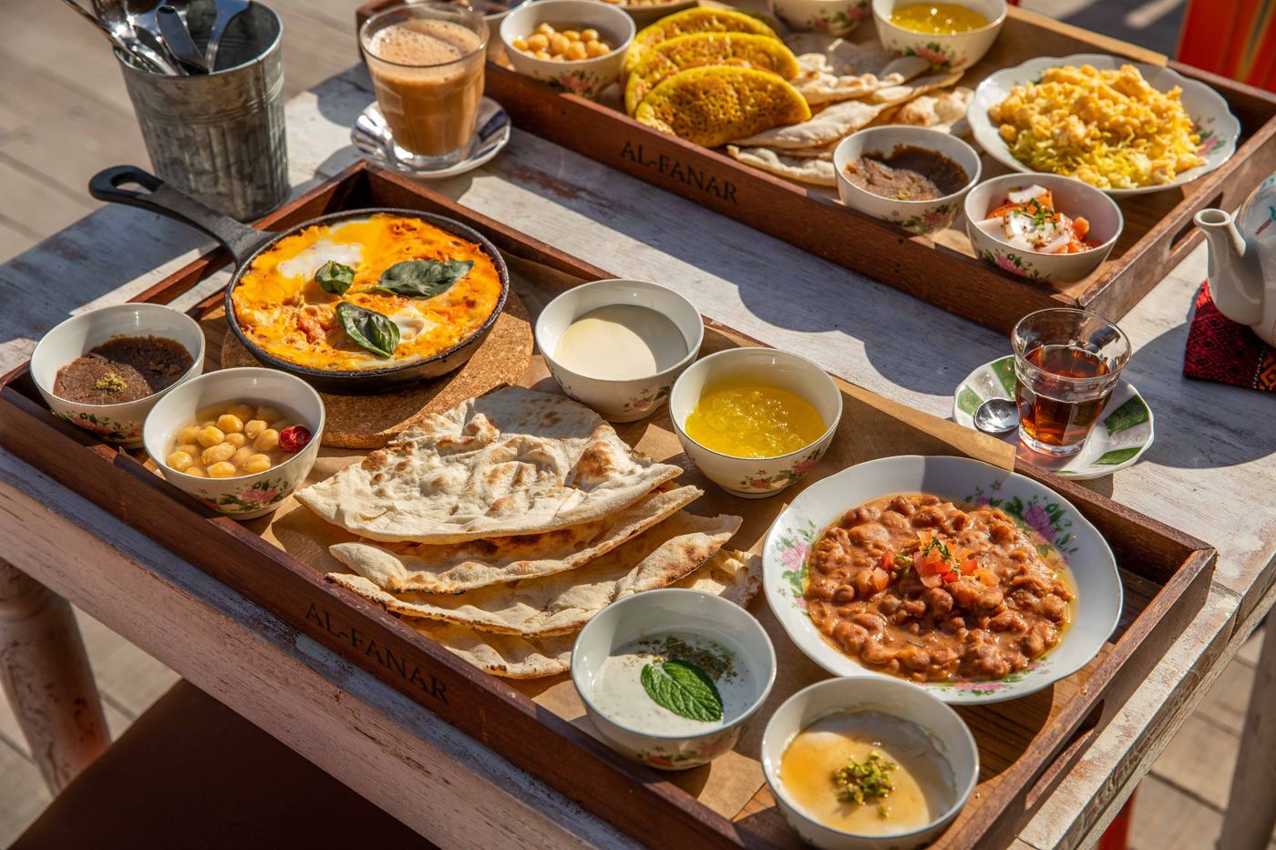 A wooden tray of Emirati breakfast dishes, bread and karak, on an outdoor table