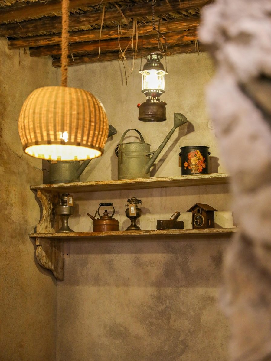 A woven lamp above a wooden shelf of old copper kettles and watering cans